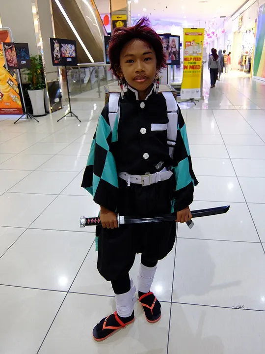 A child cosplayer with dark red hair in a green and black checkered haori, holding a sheathed sword and carrying a box on the back in a shopping mall.