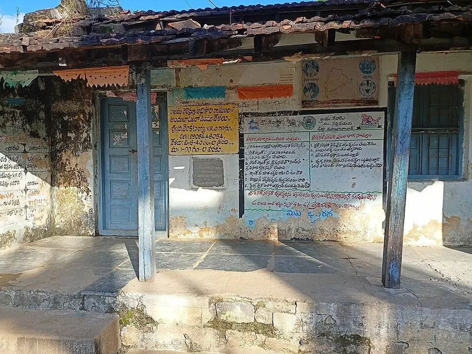 A worn village office building with a tiled roof, blue door and windows, and notice boards in a regional script on the wall.