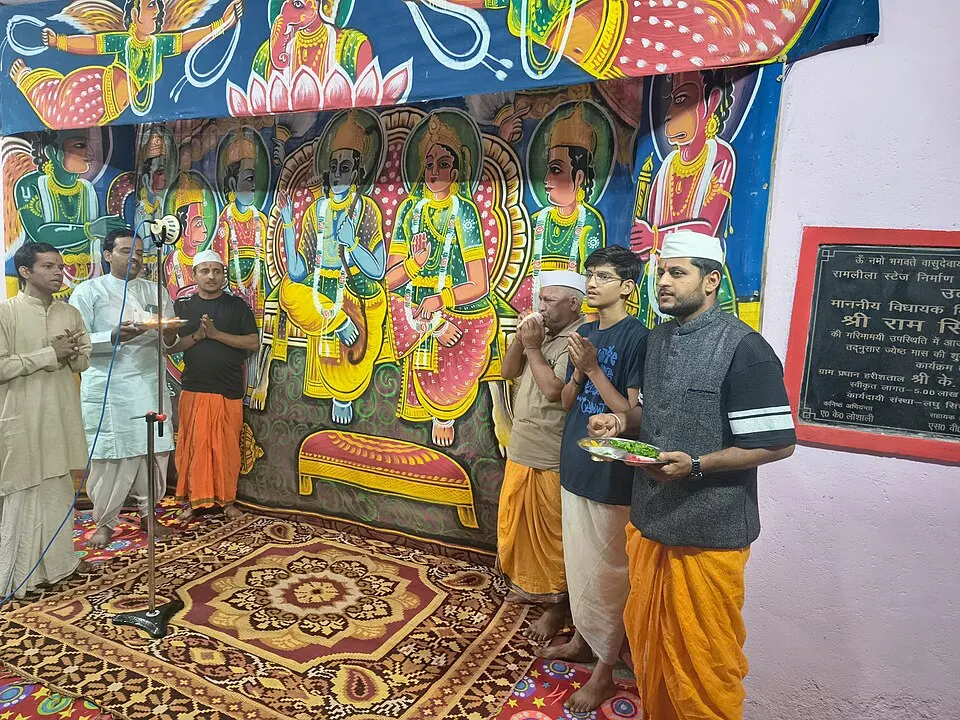Men in dhotis offer a lamp in front of a painted stage backdrop of a crowned royal couple and attendants.
