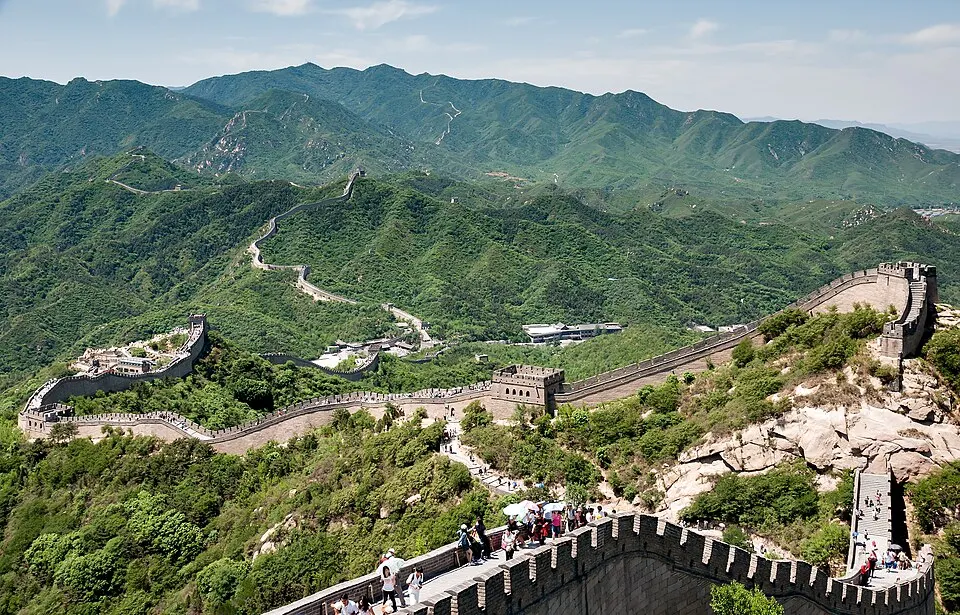 A stone wall with watchtowers winding along green mountain ridges, with a few visitors on the walkway.