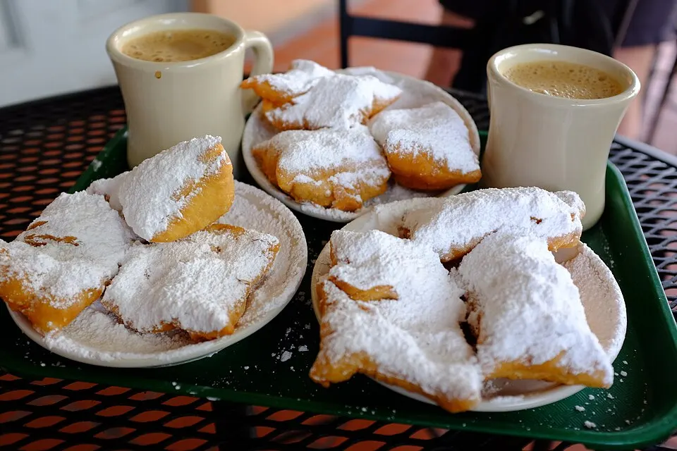 Square doughnuts covered in powdered sugar on plates beside two mugs of milky coffee on a tray.