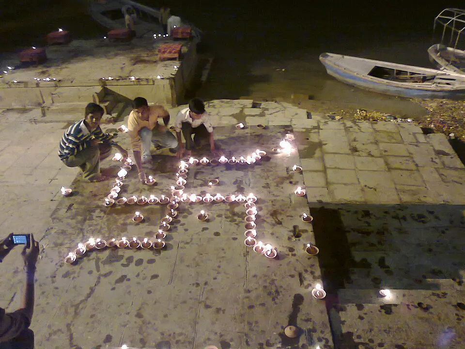 Three young men lighting small clay lamps arranged in a swastika shape on stone steps beside a river at night.
