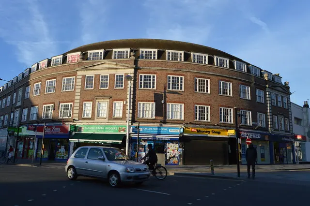 A curved brick building with shops along the ground floor on a town street, with a car and a cyclist passing.