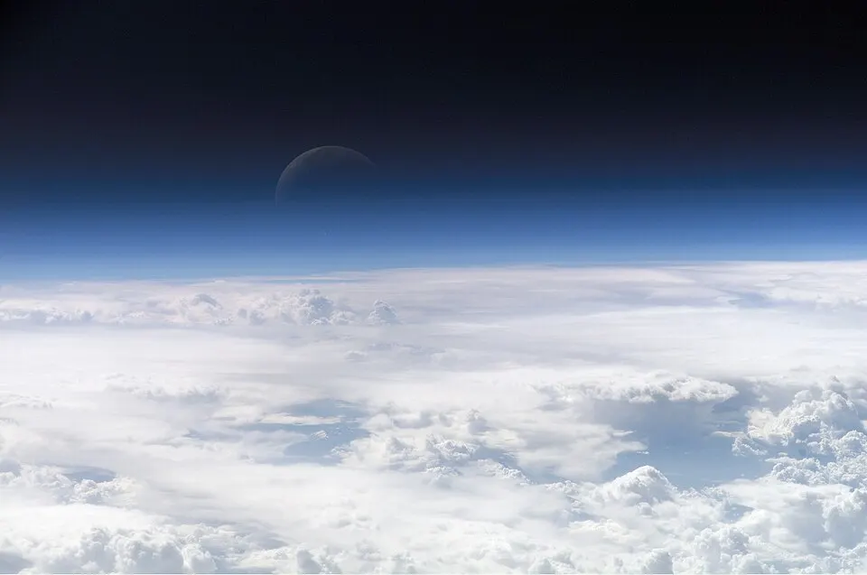 A dark crescent moon above a thin blue band at the edge of the atmosphere over a sea of white clouds.