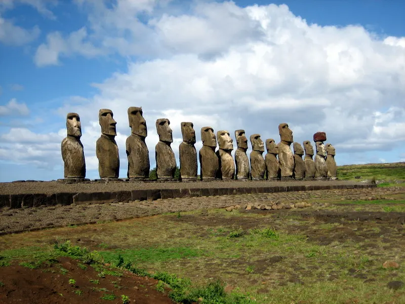 A long row of huge stone statues with heavy brows standing on a grassy platform under a cloudy blue sky.