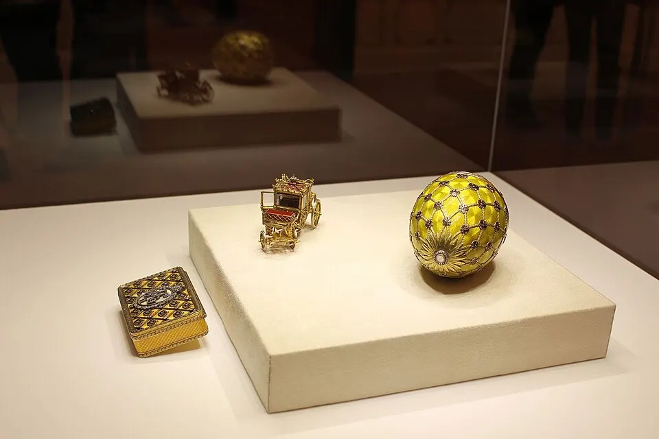 A small gold coach and a yellow enamelled jewelled egg displayed on a cream block in a glass case.