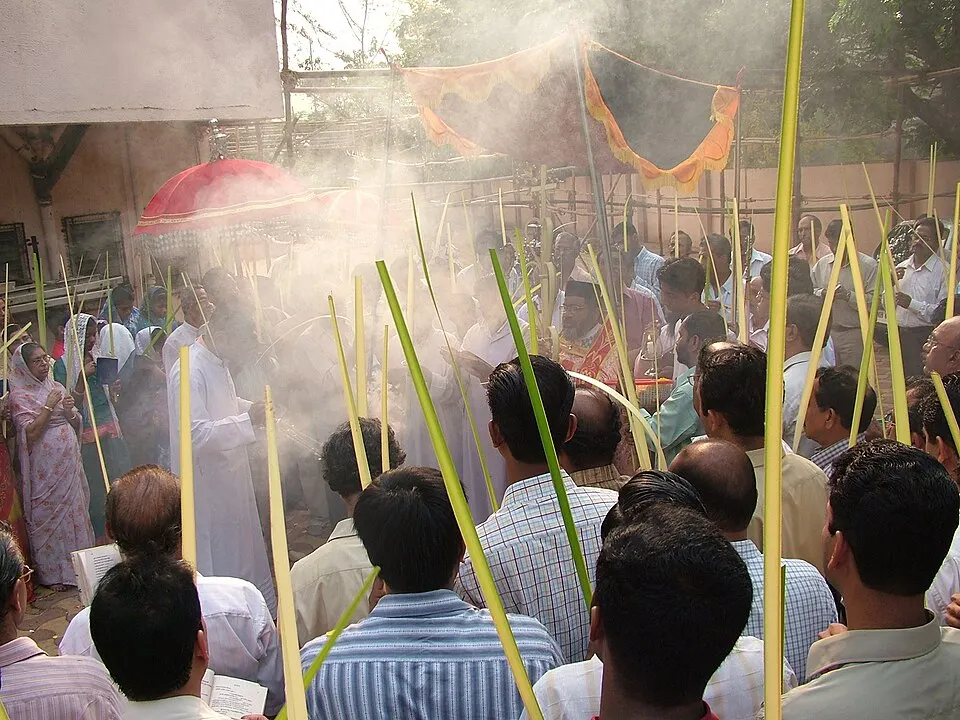 A crowd of people holding long green palm fronds in a smoky outdoor gathering, with a red umbrella and a canopy in the background.