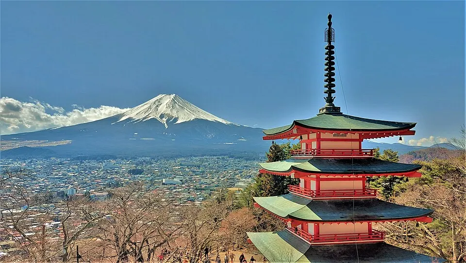 A red five-storey pagoda on a hillside above a town, with a snow-capped volcano rising in the distance.