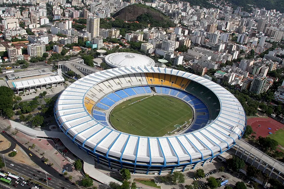An aerial view of an oval stadium with a white roof and blue and yellow seats among tall buildings and a hill.
