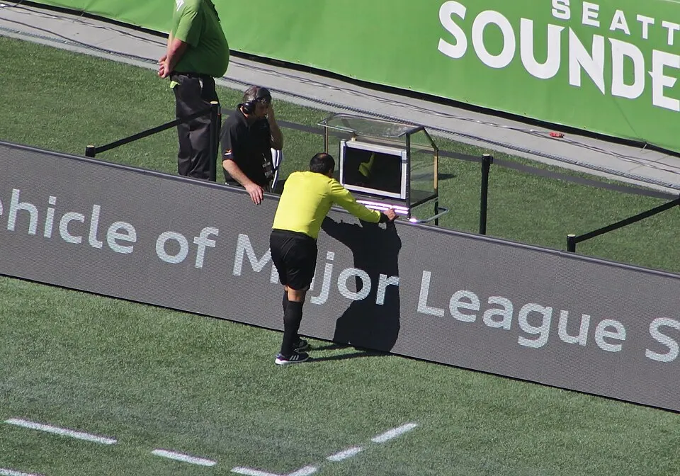 A referee in a yellow shirt leans over a small screen beside the pitch to watch a replay.