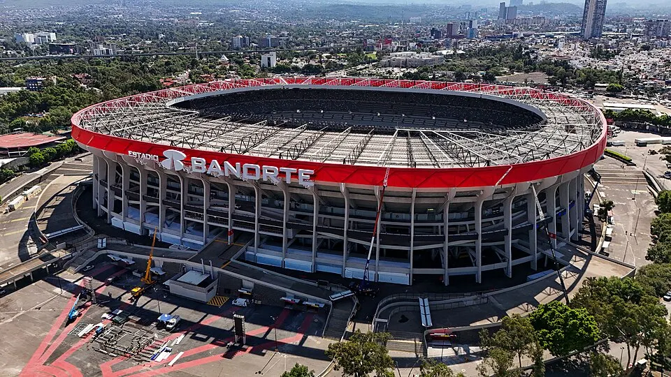 An aerial view of a huge oval stadium with a red rim and a ring of seats around a large dark bowl.
