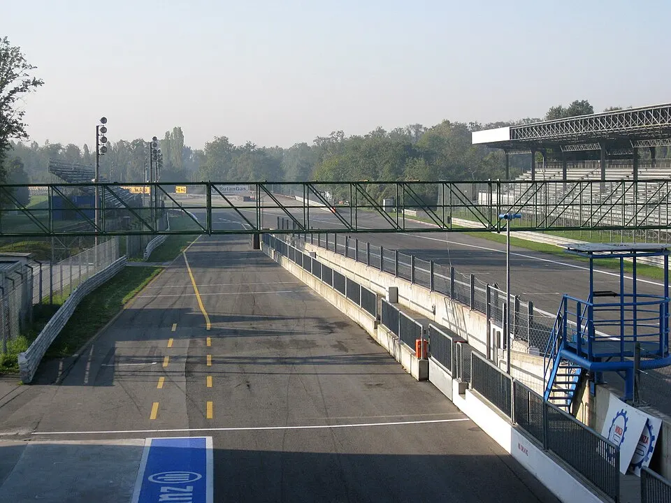 A long straight of a race track lined with trees and a pit building.