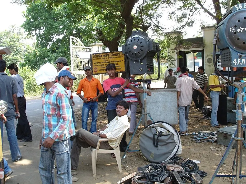 Crew and onlookers gather under trees at a film set with big studio lights, cables and a gate in the background.
