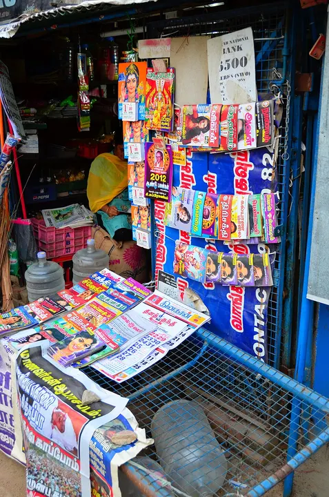 A roadside kiosk with magazines and wall calendars pinned up and stacked on a counter, a woman seated inside.