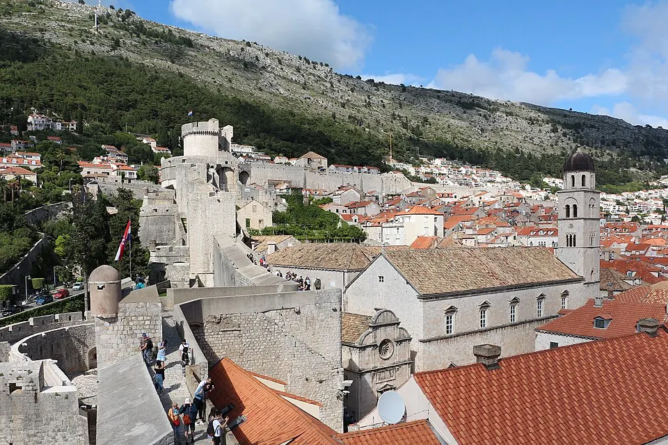 A hillside town of terracotta rooftops behind tall stone walls and towers, with a bell tower on the right.