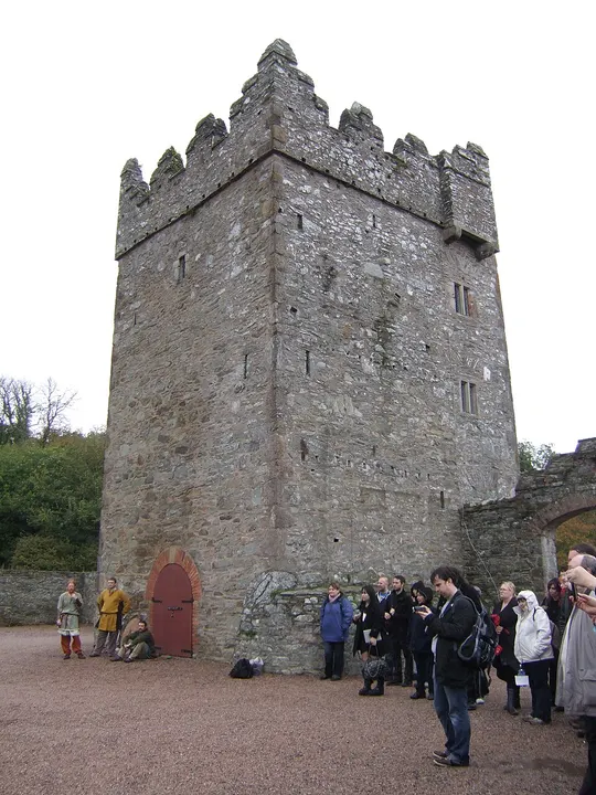 A tall grey stone castle tower with a small red arched door, with visitors standing in the courtyard.