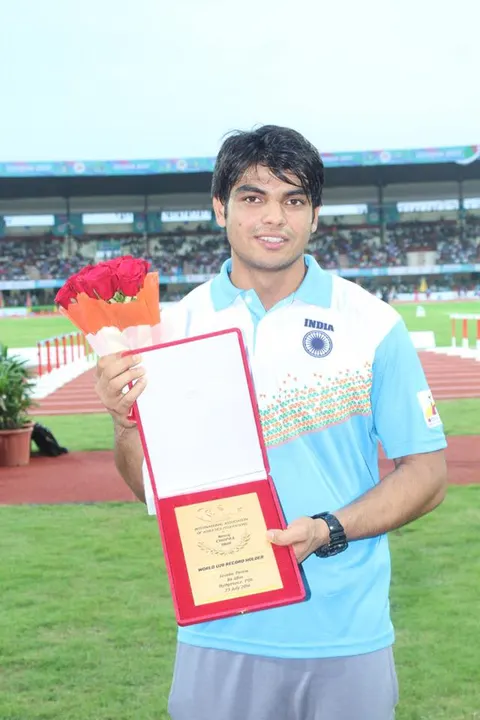 A smiling young man in a light blue sports shirt holding a framed certificate and a bouquet of red roses in a stadium.