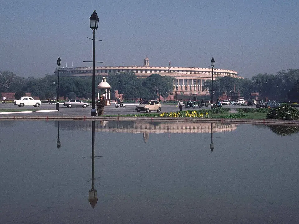 A large circular colonnaded building with a flag above it, behind lawns and flower beds.
