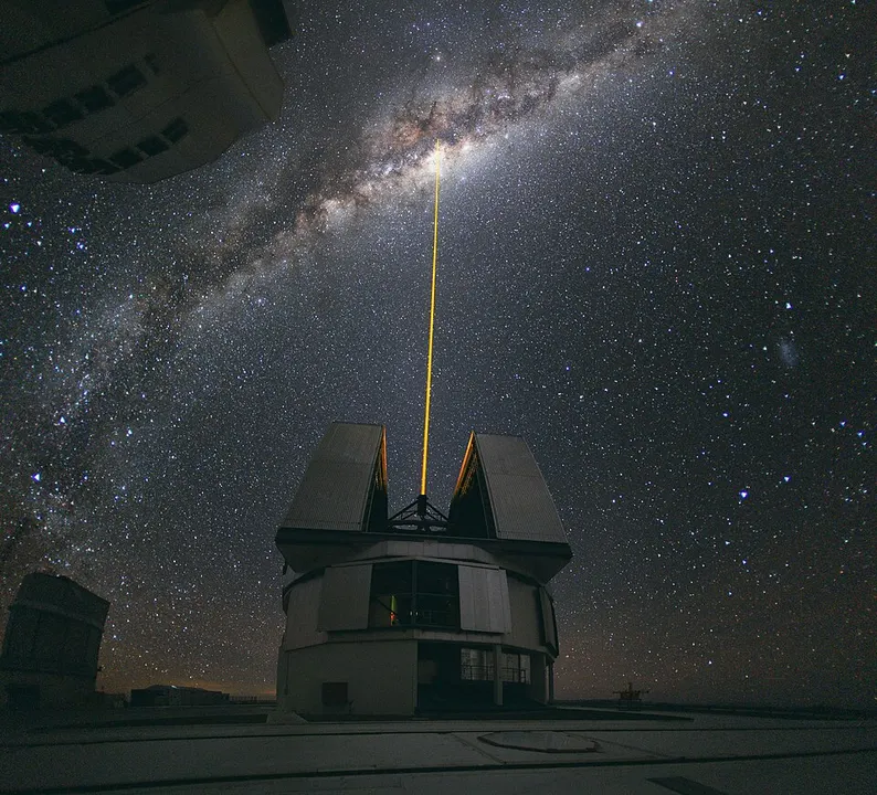 A band of countless stars stretching across a night sky above a domed observatory with a yellow beam shooting up.