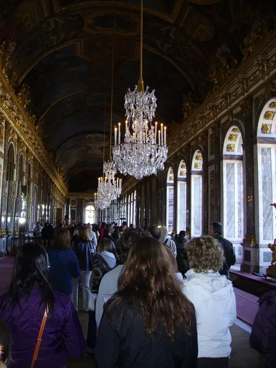 A long grand hall with a painted arched ceiling, crystal chandeliers and tall arched windows, full of visitors.