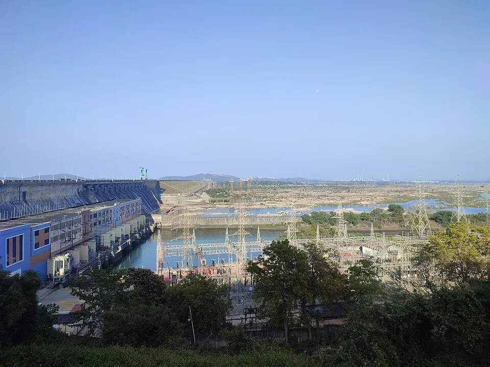 A long concrete dam wall beside a power station and electricity pylons, with water and hills beyond.