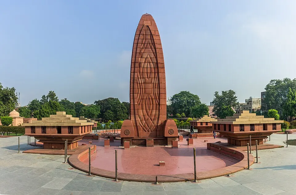 A tall red sandstone monument with carved patterns in a paved garden with small buildings around it.