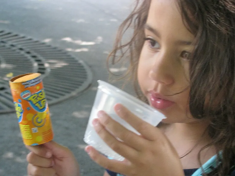 A young girl drinking from a small cup while holding an orange frozen treat.