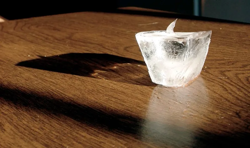 A small clear block of ice sitting in a patch of sunlight on a wooden table.