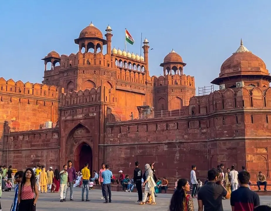 A huge red sandstone fort with domed towers, a flag on top and visitors walking in front.