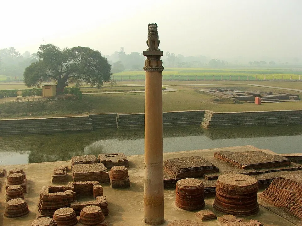 A tall sandstone pillar topped by a lion statue, standing beside a pond and brick ruins.