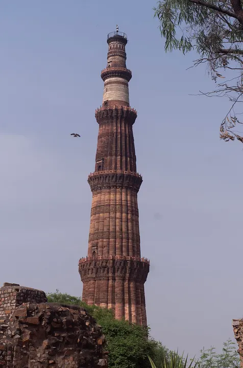 A tall tapering tower of red sandstone with rings of balconies, rising above ruins into a blue sky.