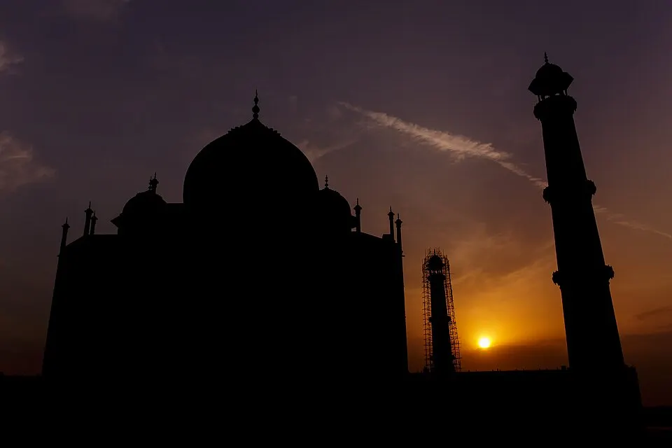 The dark silhouette of a domed building with tall minarets against an orange sunset sky.