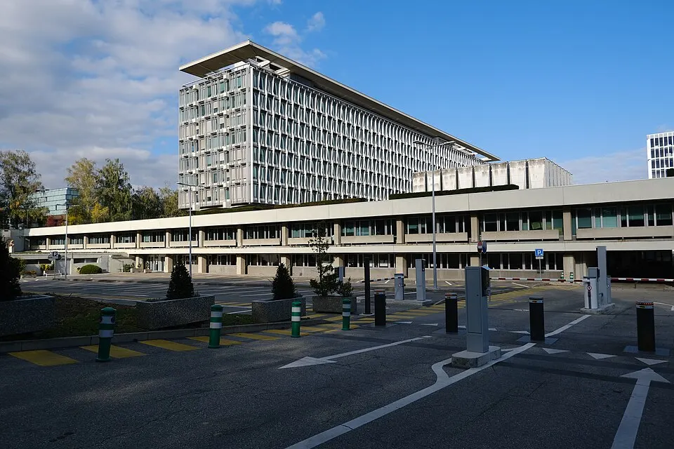 A tall, narrow modern office block with a long low building in front and a car park.