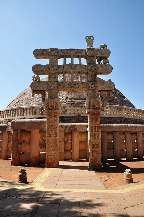 A carved stone gateway with elaborate beams in front of a large brick dome under a clear blue sky.