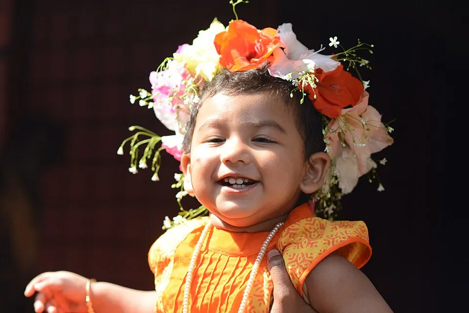 A smiling toddler wearing an orange dress and a crown of colourful flowers.