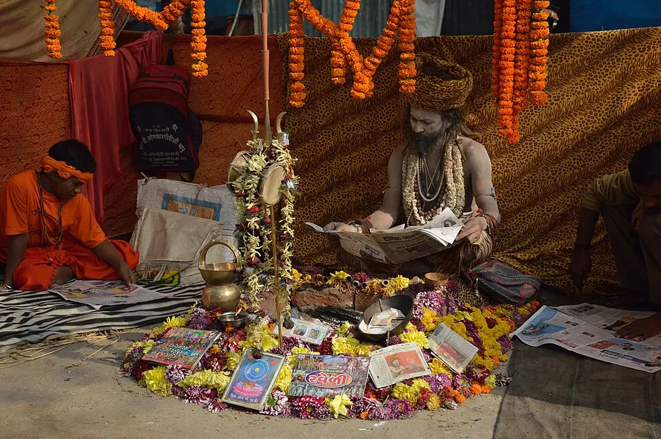 A bearded holy man in a turban sits reading a newspaper among flower garlands and printed pages spread on the ground.