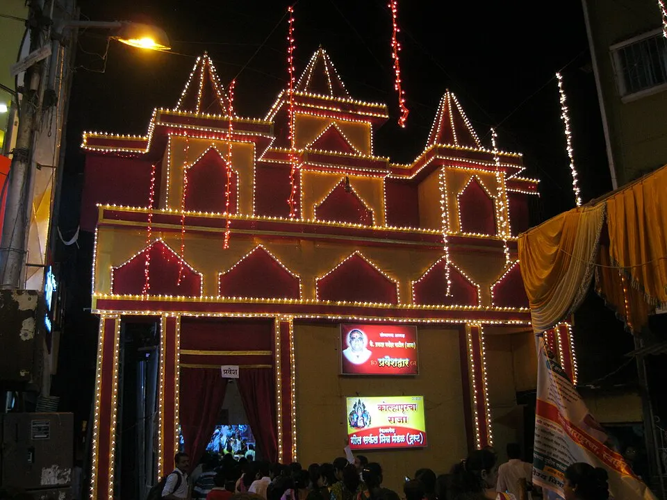 A night-time festival pandal outlined in strings of lights, with a lit entrance and a crowd waiting outside.