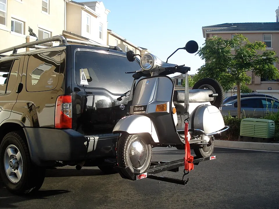A silver vintage scooter with a spare wheel is strapped to a rack at the back of a dark SUV in a car park.