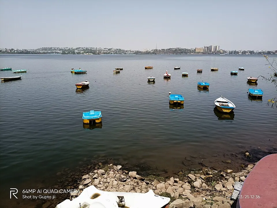 Small paddle boats and rowing boats floating on a wide, calm lake with a hazy city skyline on the far shore.