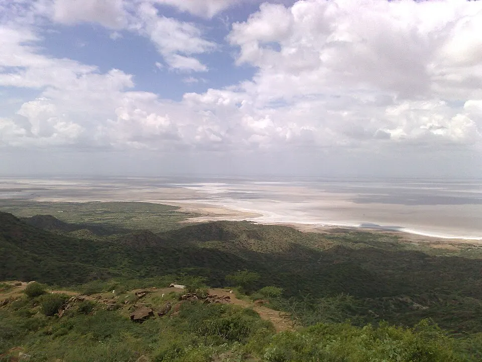 A wide view from green hills over a flat, pale salt plain stretching to the horizon under big clouds.
