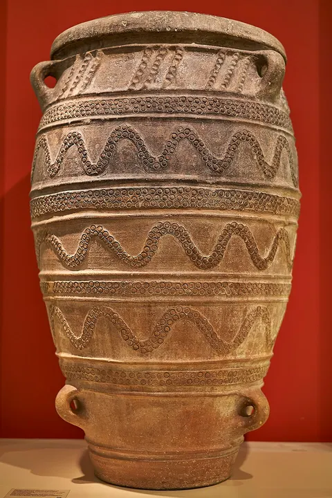 A tall ancient clay storage jar with two handles and bands of raised wavy decoration, against a red wall.