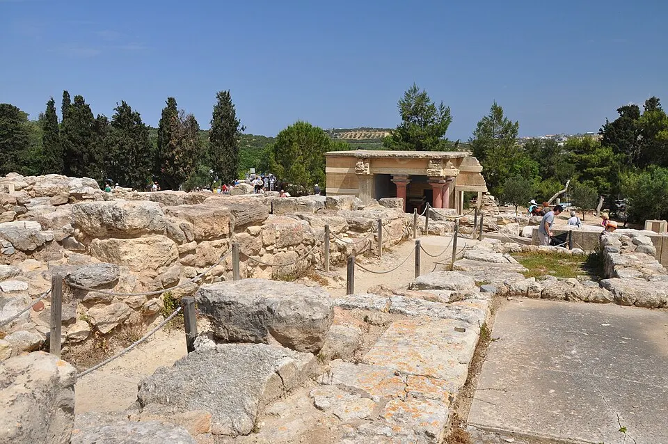 Excavated stone ruins with rope barriers and a reconstructed building with red columns among cypress trees.