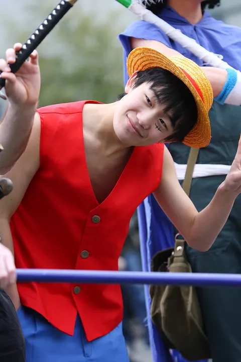 A smiling young cosplayer in a straw hat, red open vest and blue shorts at a convention.