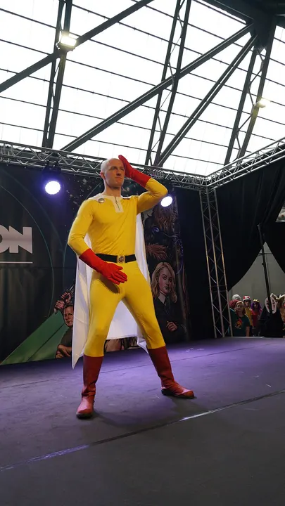 A bald-capped cosplayer in a yellow bodysuit, white cape, red gloves and red boots, saluting on a convention stage.