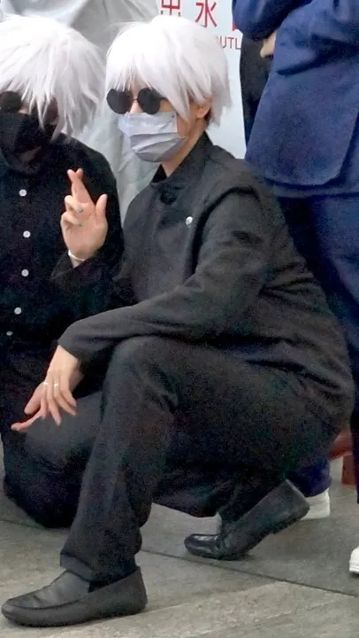 A masked cosplayer with spiky white hair and round dark glasses, in a black high-collared uniform, crouching outdoors.