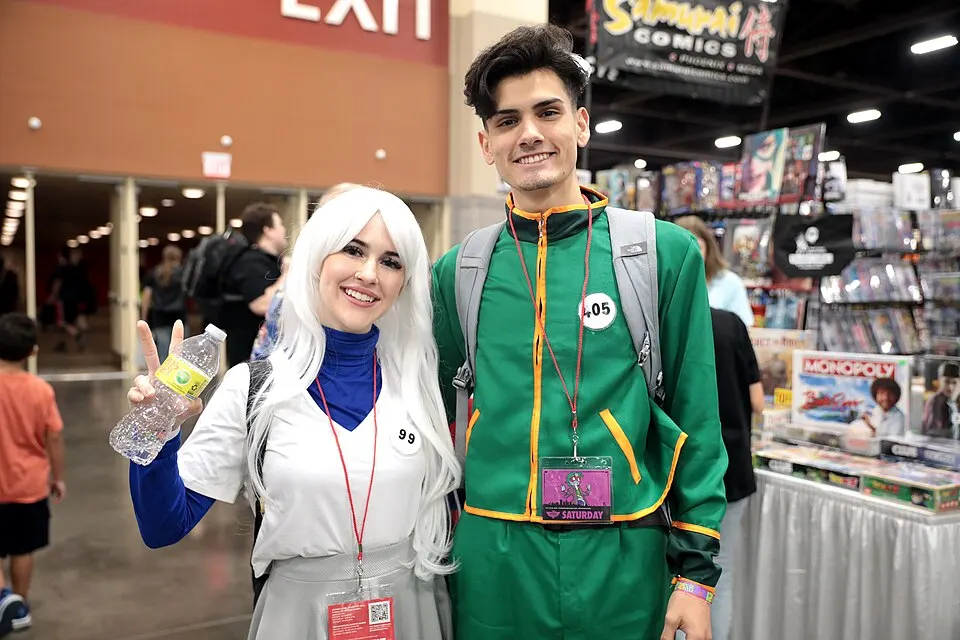 Two smiling cosplayers at a convention: a woman on the left with long white hair and a blue high-collared top, and a man on the right in a green and yellow tracksuit.