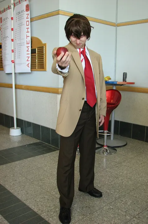 A cosplayer in a tan blazer, red tie and brown trousers holding out a red apple.
