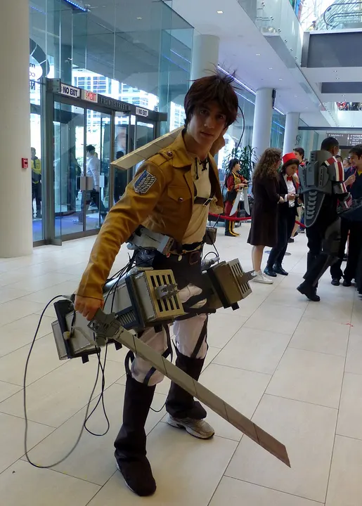 A cosplayer in a tan jacket with a crest on the sleeve, leather straps, metal gear boxes at the hips and two long blades.