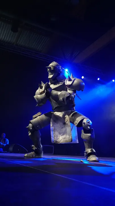 A cosplayer in a bulky grey suit of spiked armour crouching on a dimly lit stage with blue lights.