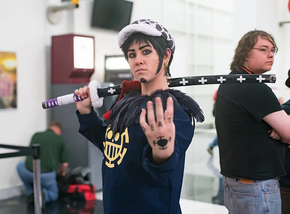A cosplayer in a spotted white hat and dark blue hoodie with a yellow emblem, a goatee and a tattooed hand held up, a long sword over the shoulder.